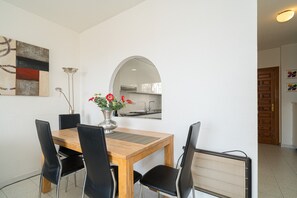 A dining area with a wooden table, black chairs, a vase with flowers, a mirror, and a kitchen visible through an archway.