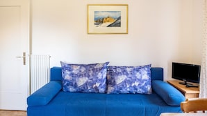 Ein Zimmer mit einem blauen Sofa, einem gerahmten Bild an der Wand, einem Heizkörper, einem Fernseher auf einem Holztisch und einer Tür.