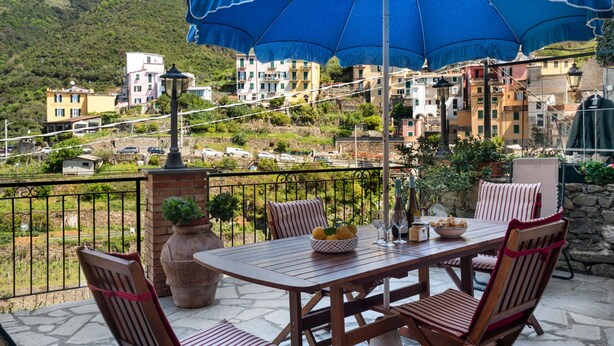 Un patio exterior con una mesa y sillas, un gran paraguas azul y vistas a un pueblo en la ladera.