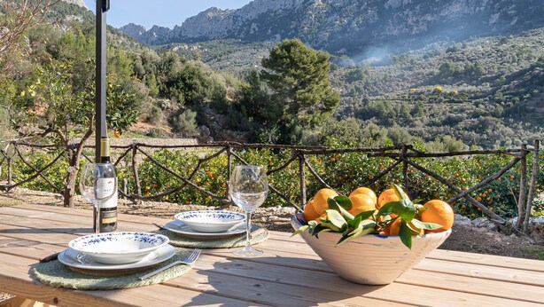 A table set outdoors with plates, wine glasses, and a bowl of oranges, overlooking a mountainous landscape.