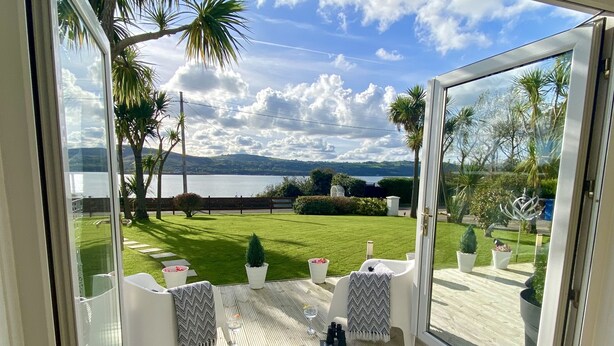 A balcony with a view of a lake, a grassy lawn, and a wooden deck. - Seaview Cottage on the Island (Islandmagee)