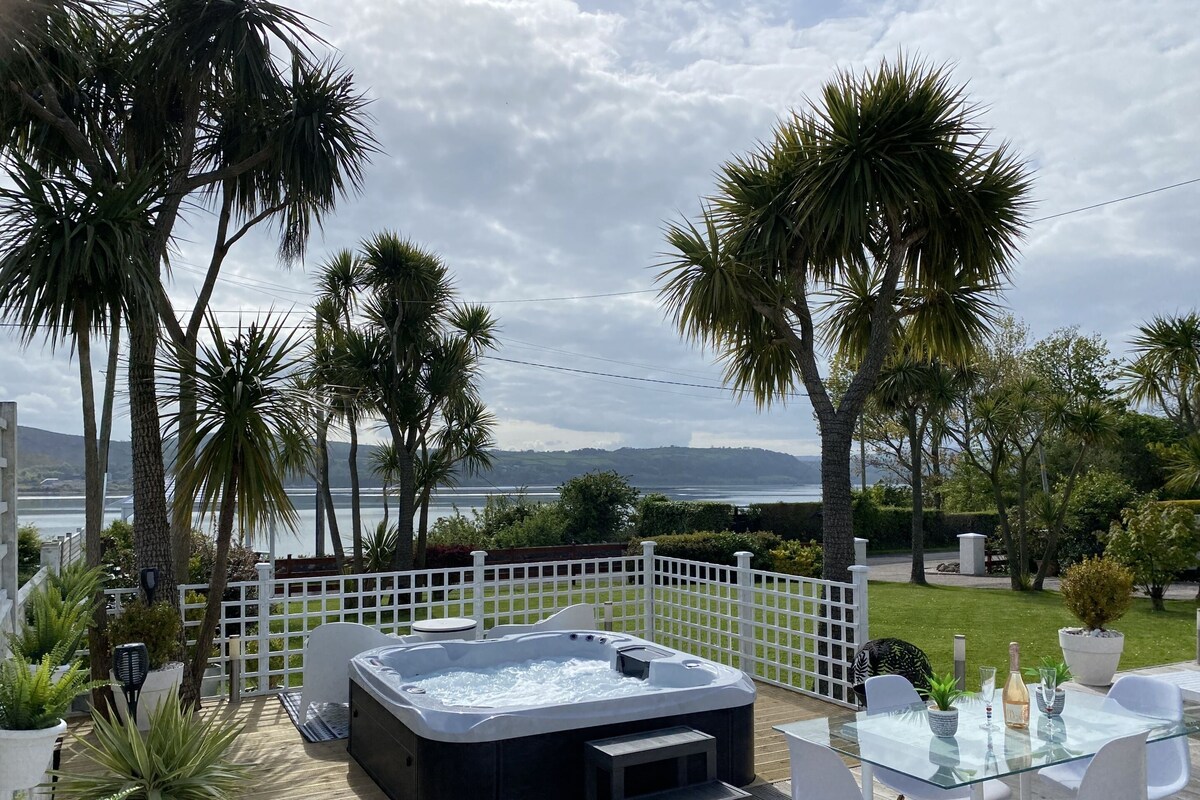 A hot tub with a glass table and chairs, surrounded by palm trees and a view of the sea.