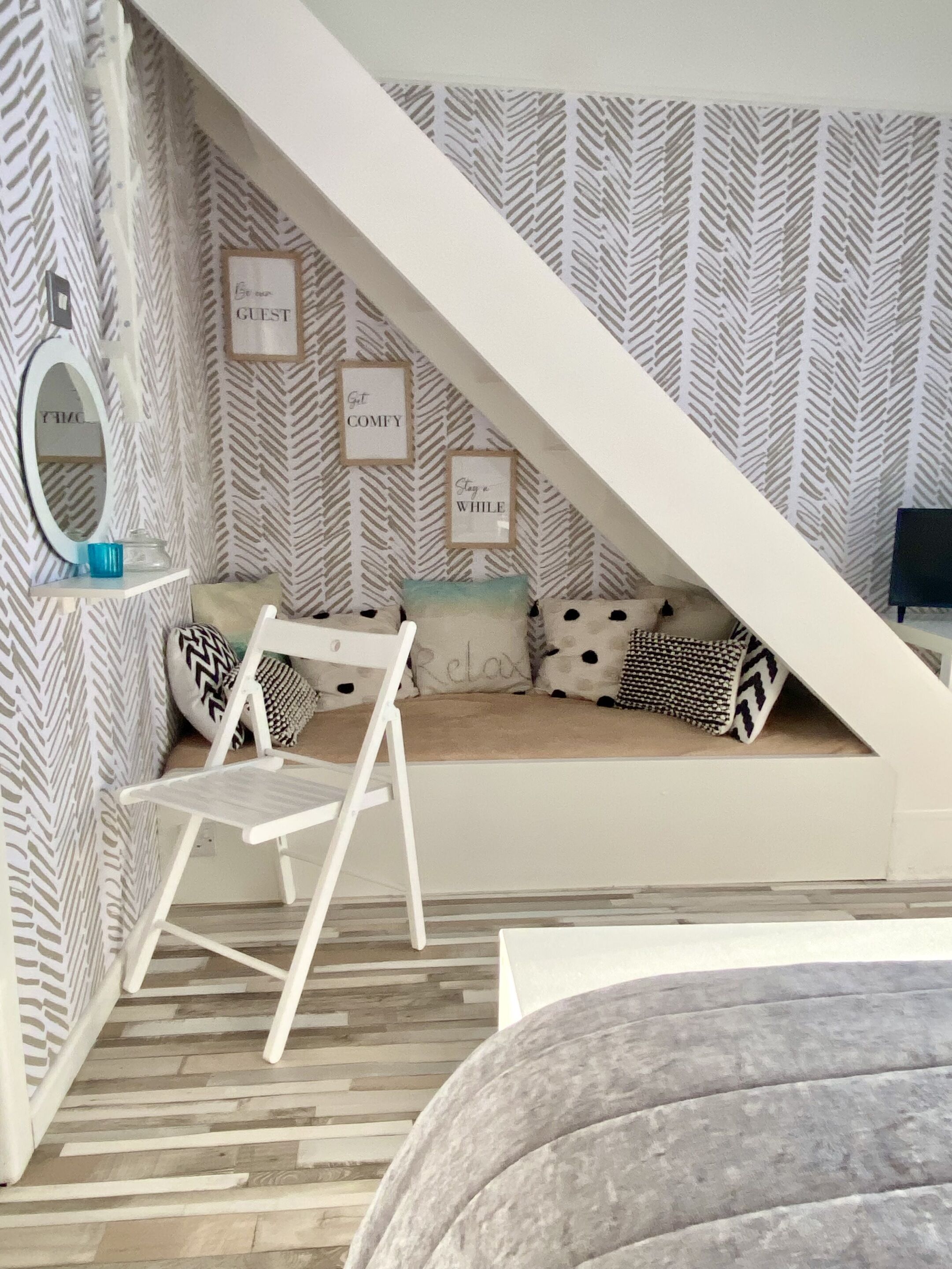 Un dormitorio acogedor en el ático, con techo inclinado, una escalera blanca, un banco acolchado y una pequeña mesa con un objeto azul.