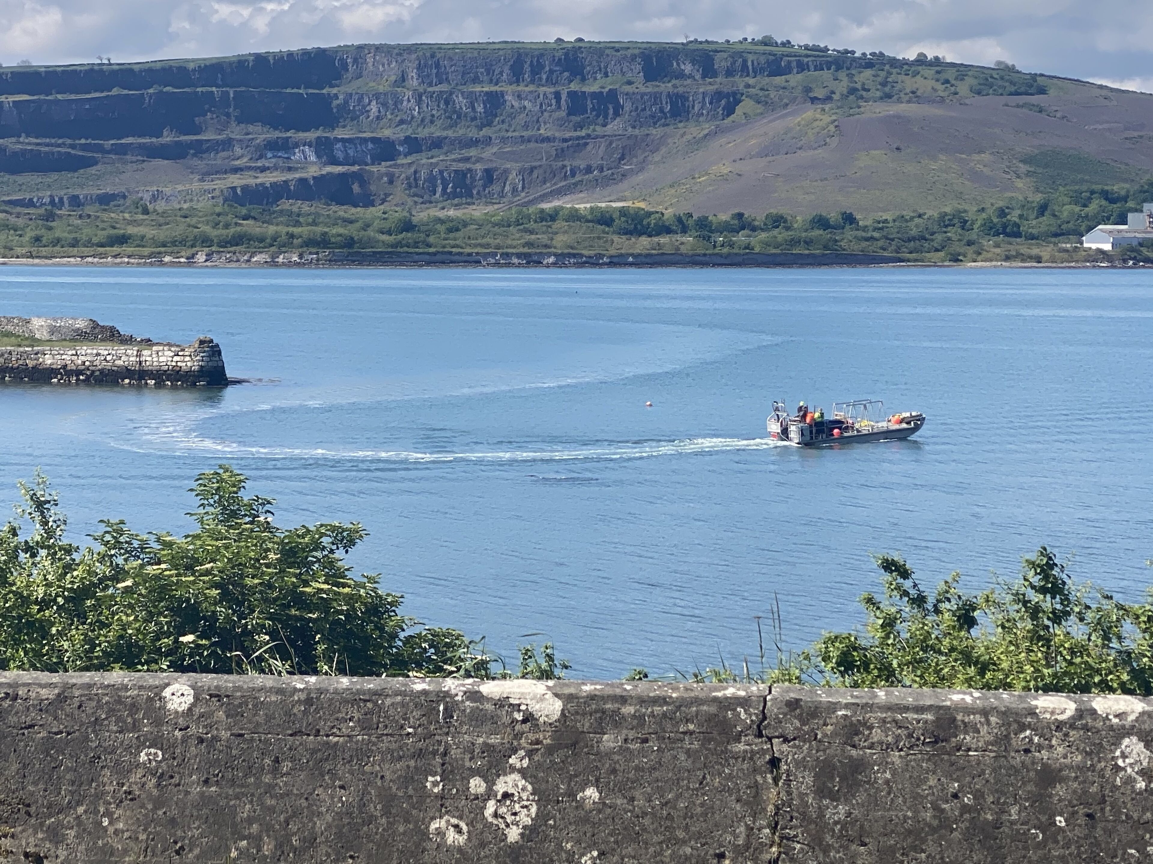 A boat on a calm body of water with a stone structure on the left and a hilly landscape in the background.