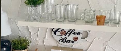 Um balcão de café com prateleiras exibindo copos, uma máquina de café e uma placa com a inscrição 'Coffee Bar'.