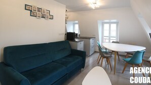 Un salon moderne avec un canapé de couleur turquoise, un coin repas avec une table blanche et des chaises bleues, et une cuisine équipée d'une machine à café noire.