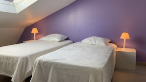 Une chambre dotée de deux lits, chacun recouvert d’une couvre-lit blanche et agrémenté d’un oreiller assorti. De chaque côté des lits, on trouve des tables de chevet surmontées de lampes. La chambre a un plafond en pente avec une lucarne, et les murs sont peints en violet.