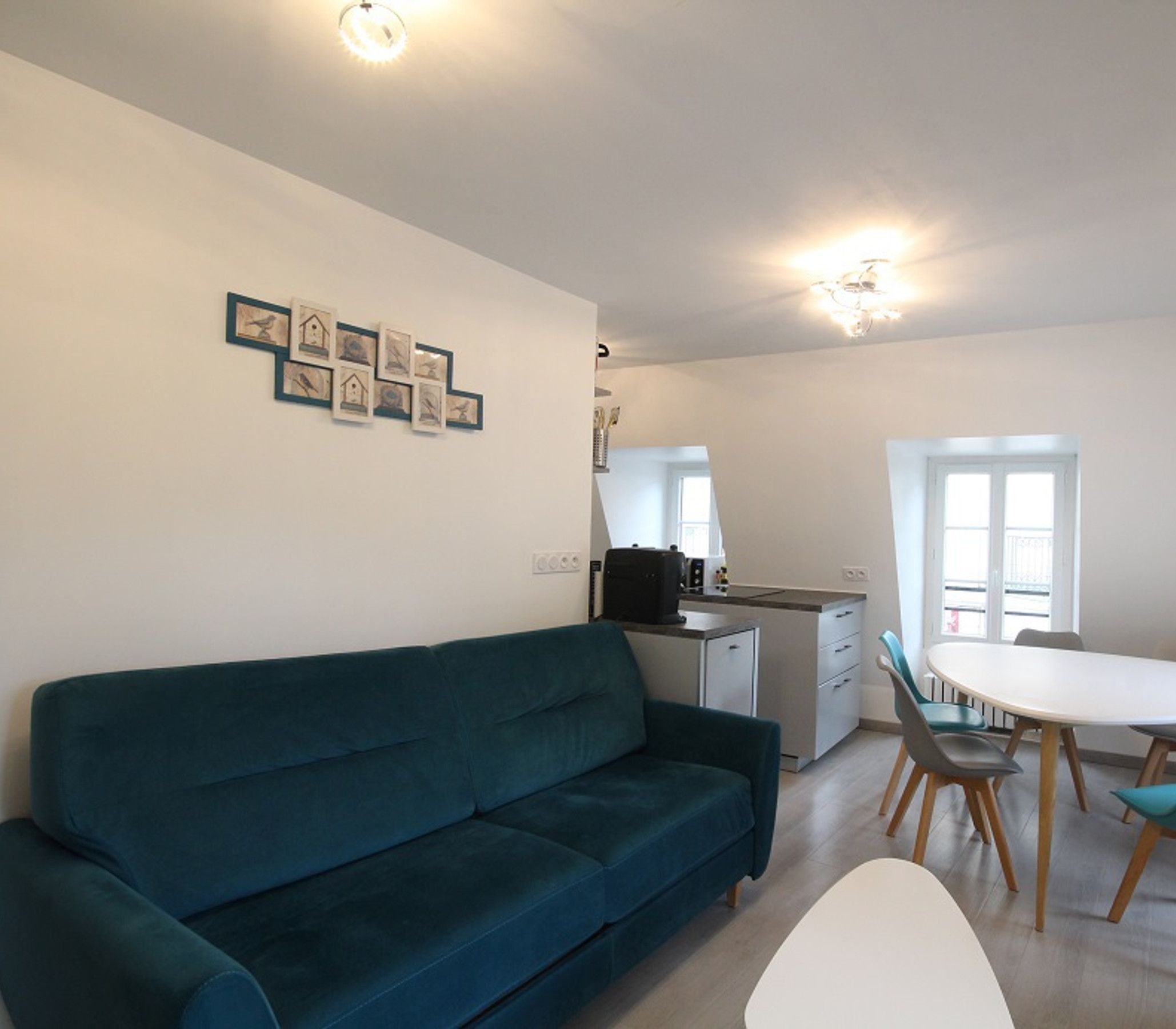 A modern living room with a teal sofa, a dining area with a white table and blue chairs, and a kitchen with a black coffee machine.