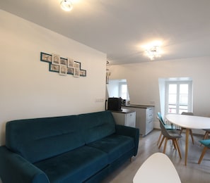 A modern living room with a teal sofa, a dining area with a white table and blue chairs, and a kitchen with a black coffee machine.