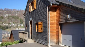 A two-story wooden house with a garage, a small porch, and a view of a residential area with houses and trees. - Mont-Dore: 4-bedroom chalet with garage and terrace (Mont-Dore)