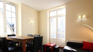 Un salón moderno con una mesa de comedor de madera, sillas negras, una mesita auxiliar roja, un sofá negro con un cojín rojo, y un ventanal que proporciona luz natural.
