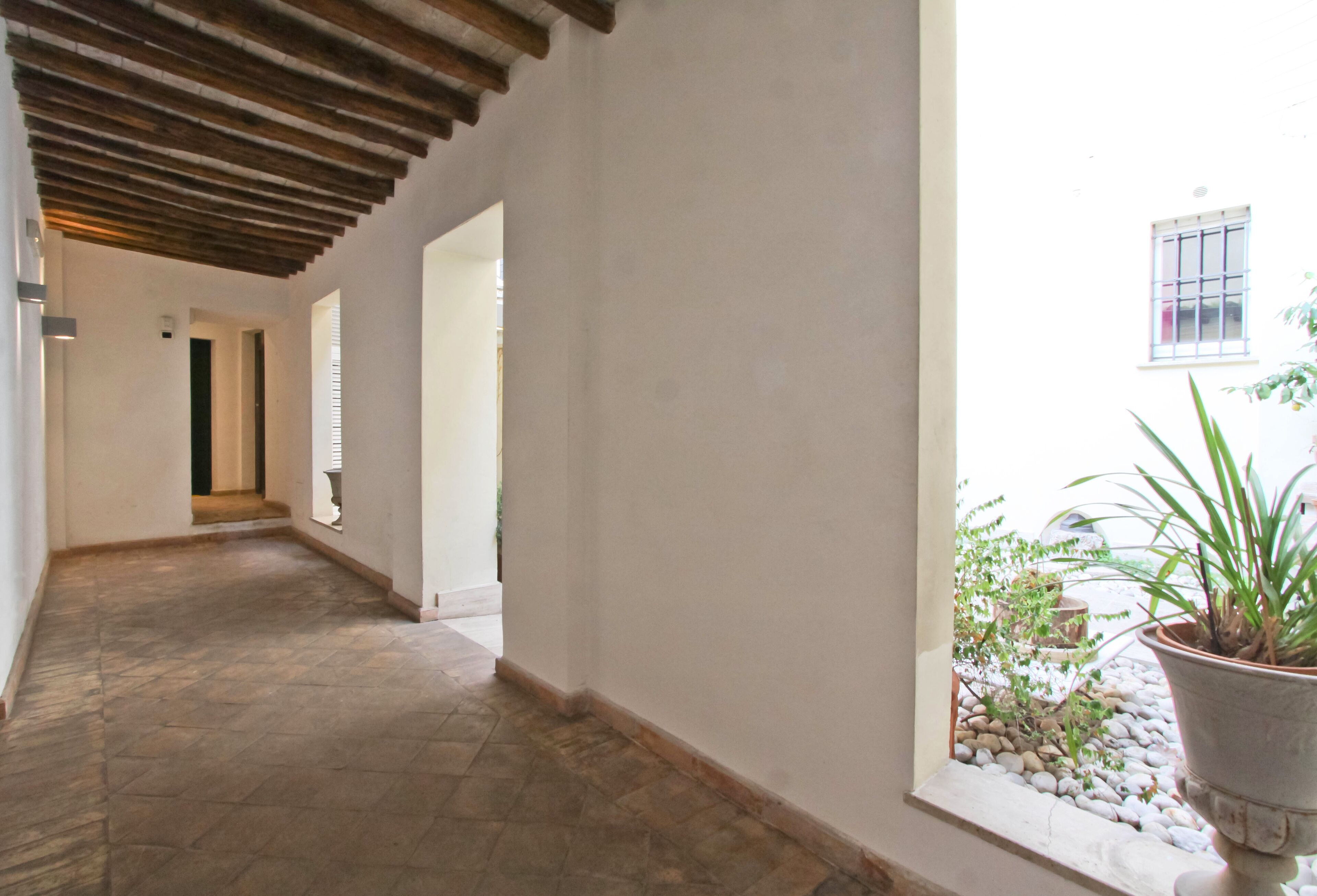A corridor with a tiled floor, wooden ceiling beams, and white walls. There is a window with a view of a garden with plants and a potted plant.