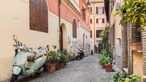 A narrow cobblestone street with a vintage scooter parked on the left, flanked by buildings with arched windows and potted plants.