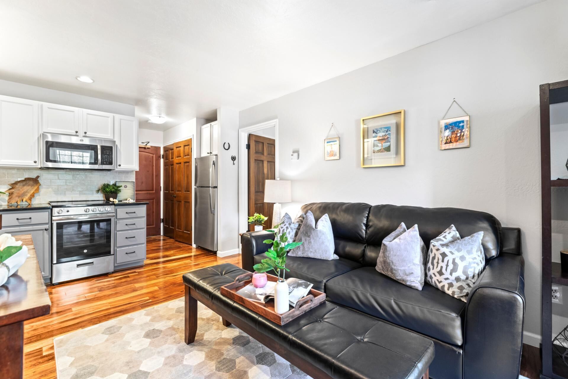 A modern kitchen with stainless steel appliances, a dining area with a wooden table and bench, and a living room with a black leather sofa and decorative pillows.
