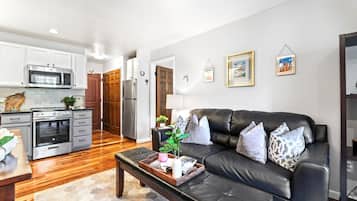 A modern kitchen with stainless steel appliances, a dining area with a wooden table and bench, and a living room with a black leather sofa and decorative pillows.