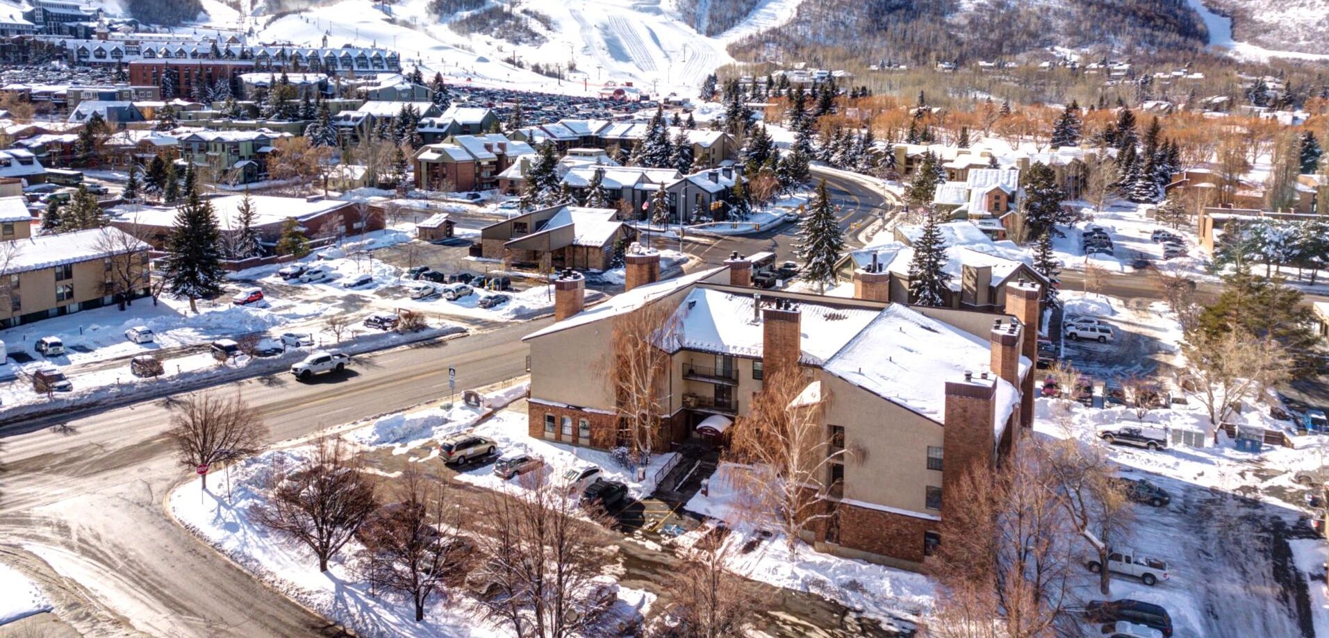 A snow-covered town with multiple buildings, a main road, and a parking area.