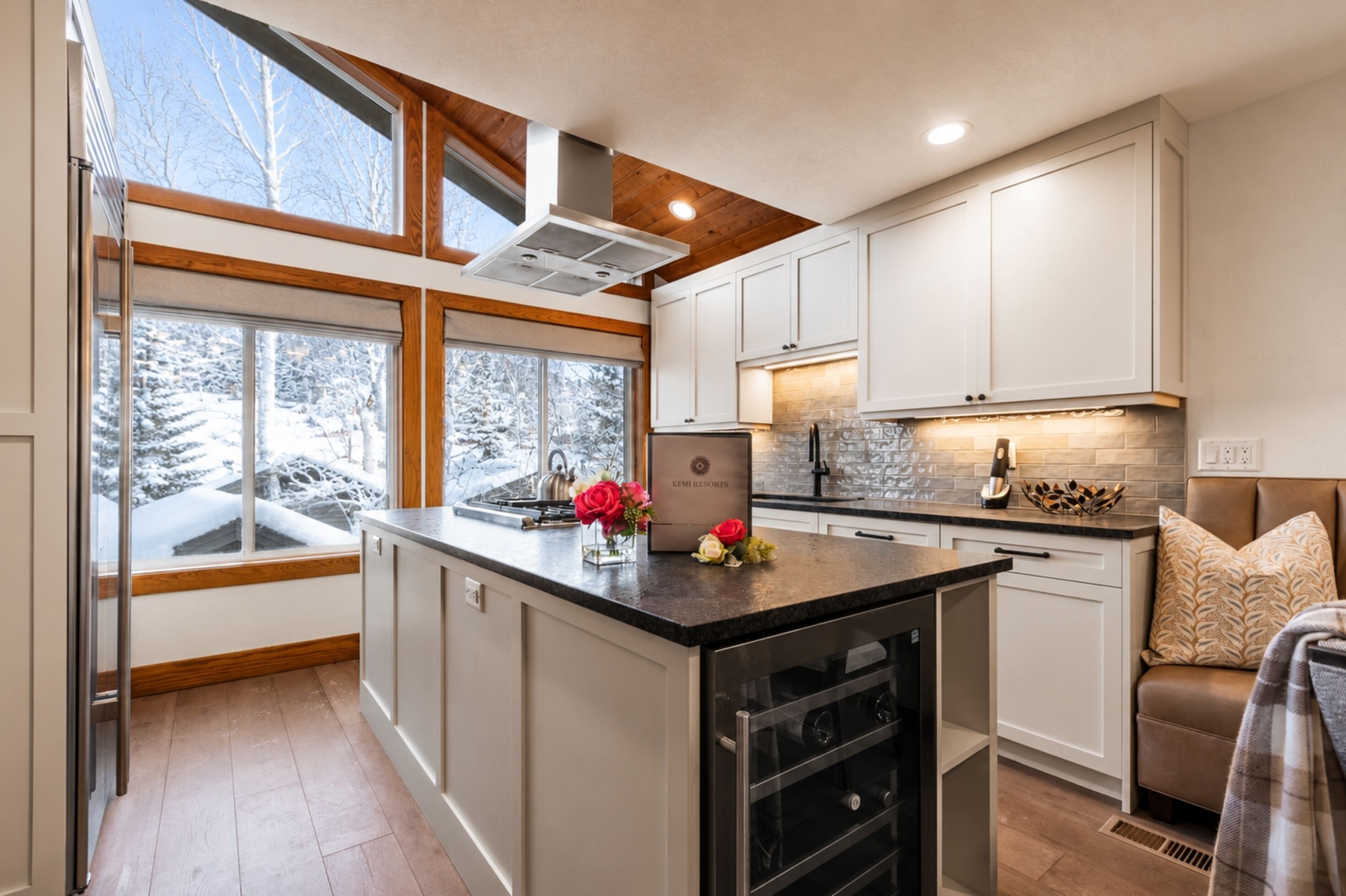 A modern kitchen with a large island, white cabinetry, and a view of snow-covered trees through large windows.