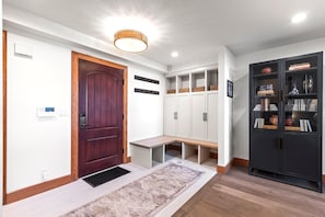 Un couloir avec une porte en bois, un banc, un meuble blanc avec des étagères et un meuble noir orné d’objets décoratifs.