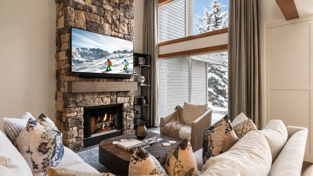 A cozy living room with a stone fireplace, a flat-screen TV, and a large window with a view of snow-covered trees. - Boulder Creek 3 Bedrooms BCC-1101 by KBM (Park City)