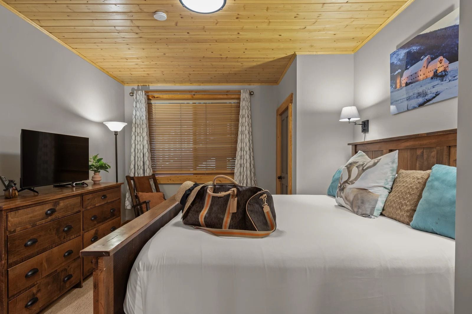 Ein Hotelzimmer mit Holzboden, einem Bett mit vielen Kissen, einem Fernseher auf einer Holzkommode und einem Fenster mit Jalousien.