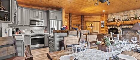 A cozy log cabin interior with a dining area set for a meal, featuring wooden furniture, a stone fireplace, and modern appliances.