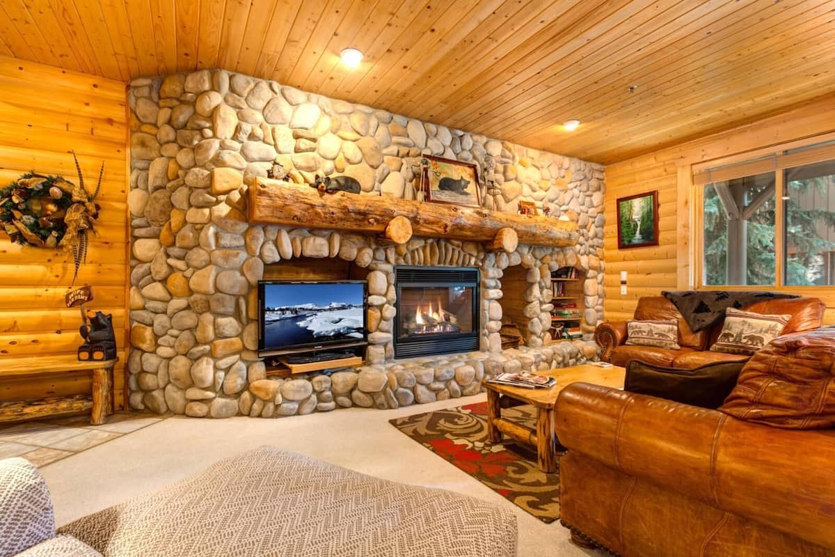 A log cabin interior with a stone fireplace, a television, a leather couch, and a bookshelf.