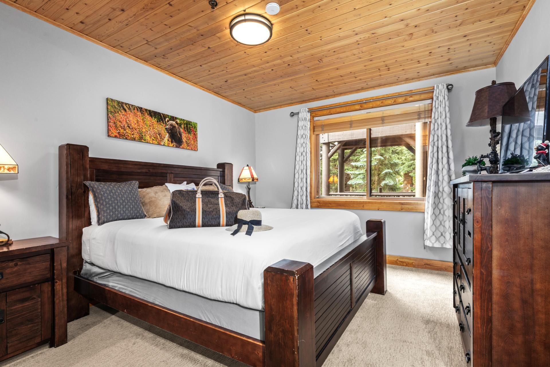 A bedroom with a wooden ceiling, a bed with white bedding, a nightstand, a lamp, a window with curtains, and a painting on the wall.