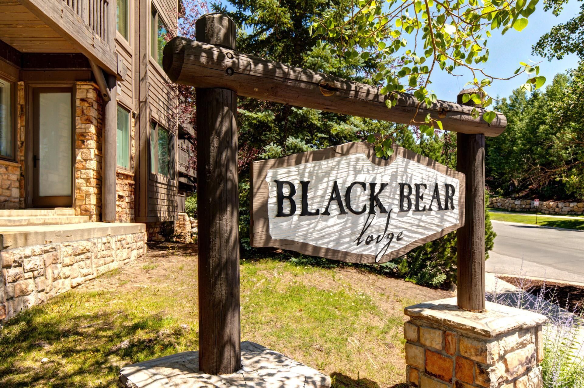 A wooden sign with "BLACK BEAR lodge" written on it, supported by a wooden post and stone base, with a stone building in the background.