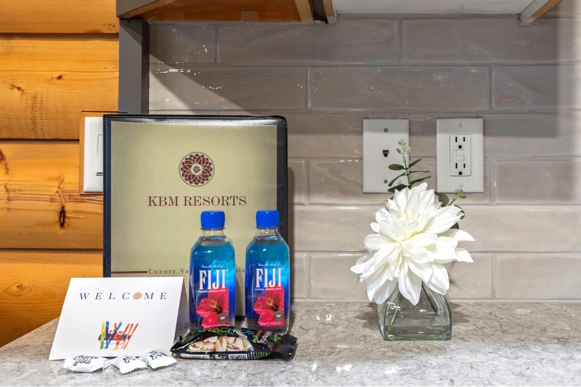 A welcome card, two bottles of Fiji water, a floral arrangement, and a gift basket with chocolates and snacks.