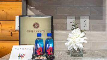 A welcome card, two bottles of Fiji water, a floral arrangement, and a gift basket with chocolates and snacks.