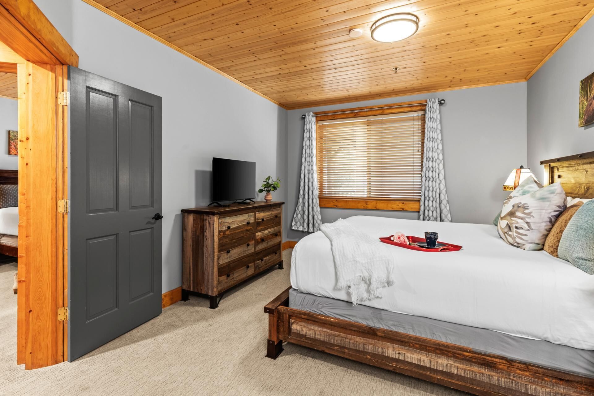 A bedroom with a wooden ceiling, a bed with a red tray, a television on a wooden dresser, and a window with blinds.