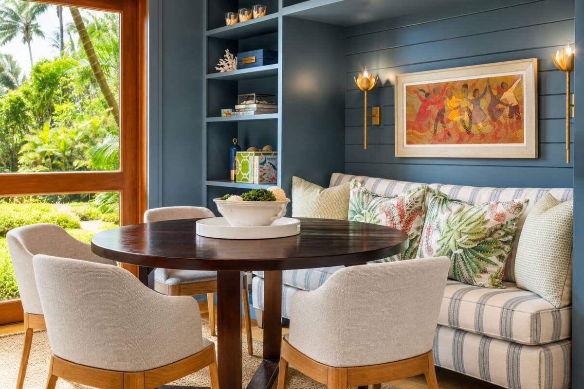 A room with a round table, four chairs, a sofa with patterned cushions, a bookshelf, and a framed artwork on the wall.