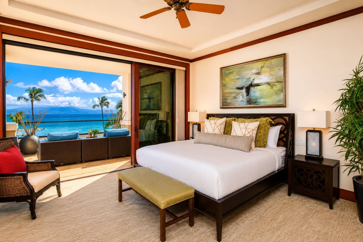 A hotel room with a large bed, a bench, a chair, a nightstand, a painting, and a view of the ocean.