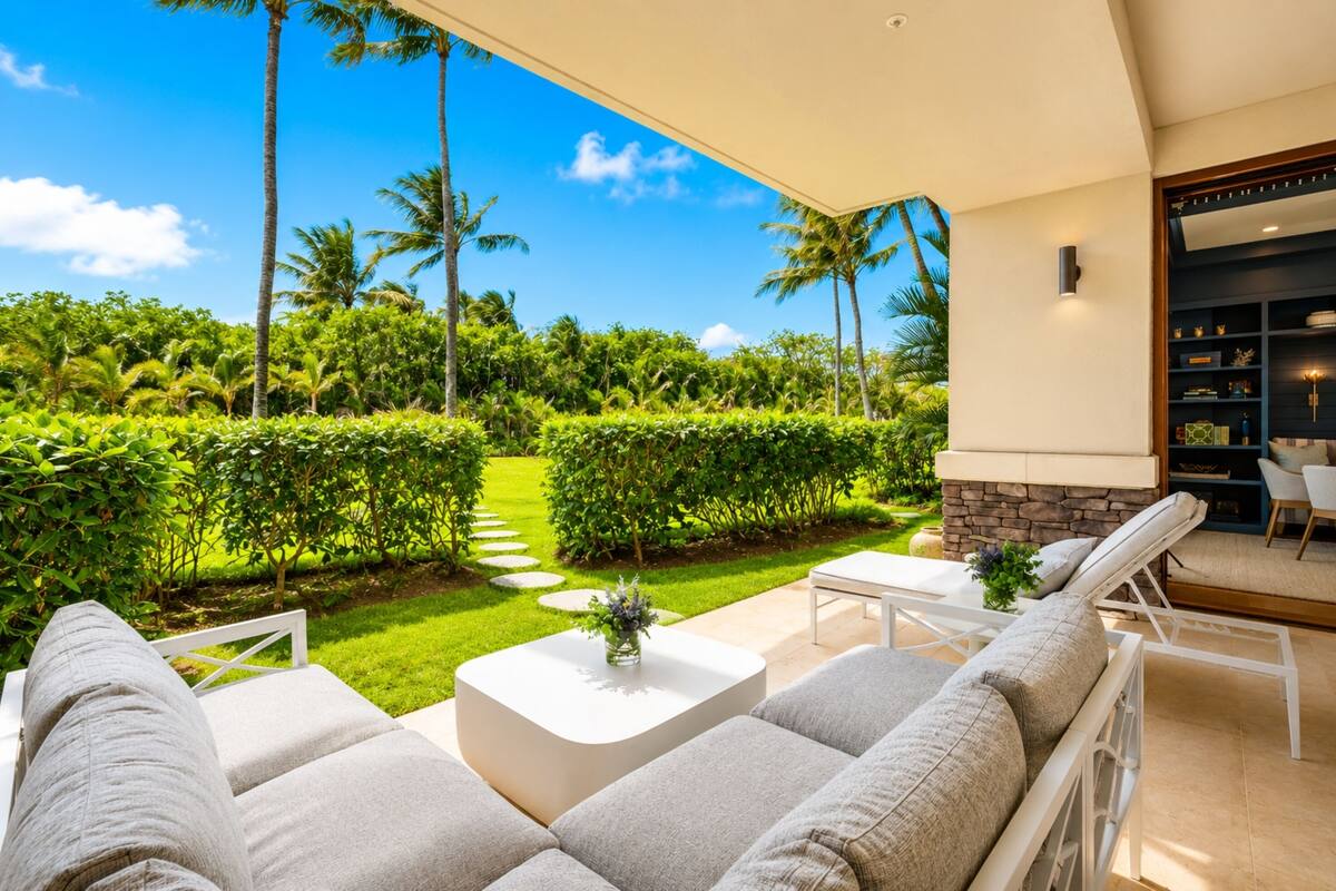 A modern outdoor seating area with a stone fireplace, white furniture, and a view of a well-maintained garden with palm trees.