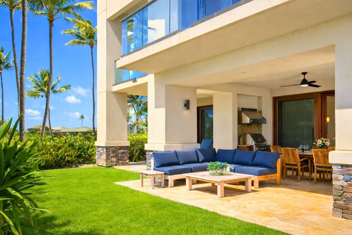 A modern outdoor patio with a sectional sofa, dining table, and chairs, surrounded by a well-maintained lawn and palm trees.