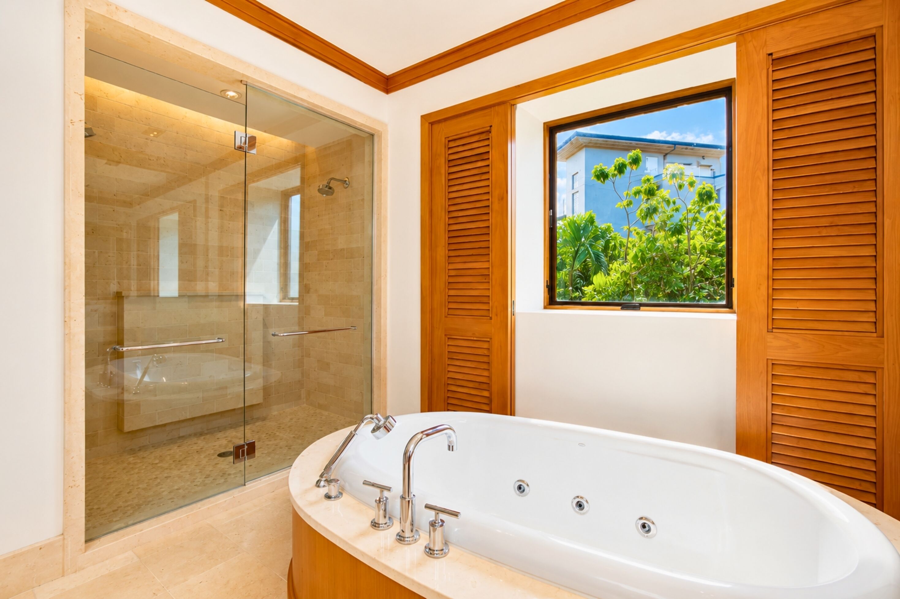 A modern bathroom with a glass-enclosed shower, a freestanding bathtub, wooden shutters, and a window with a view of greenery.