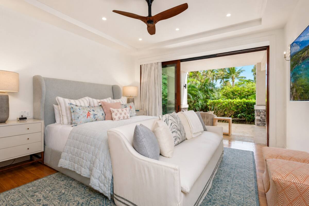 A bedroom with a bed, a sofa, a ceiling fan, a nightstand, and a window with a view of greenery.