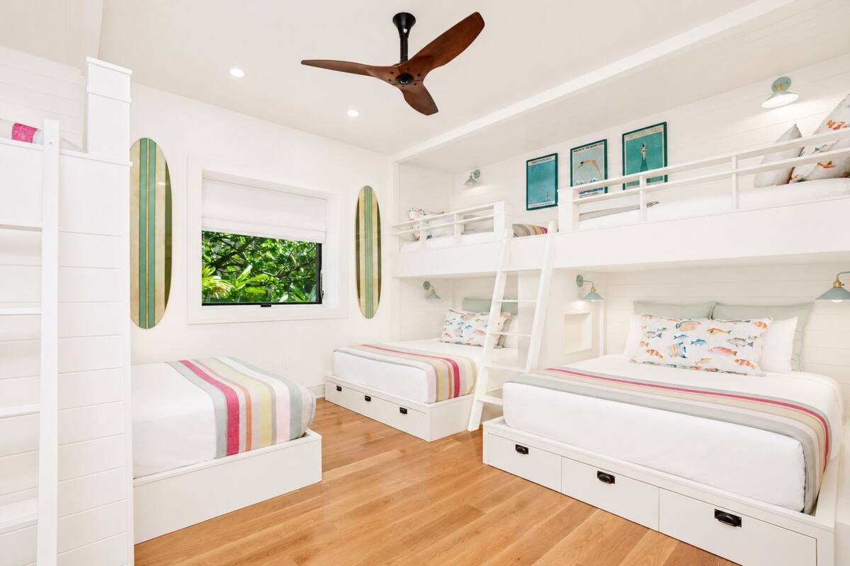 A room with bunk beds, a ceiling fan, a window with a view of greenery, and wall art.