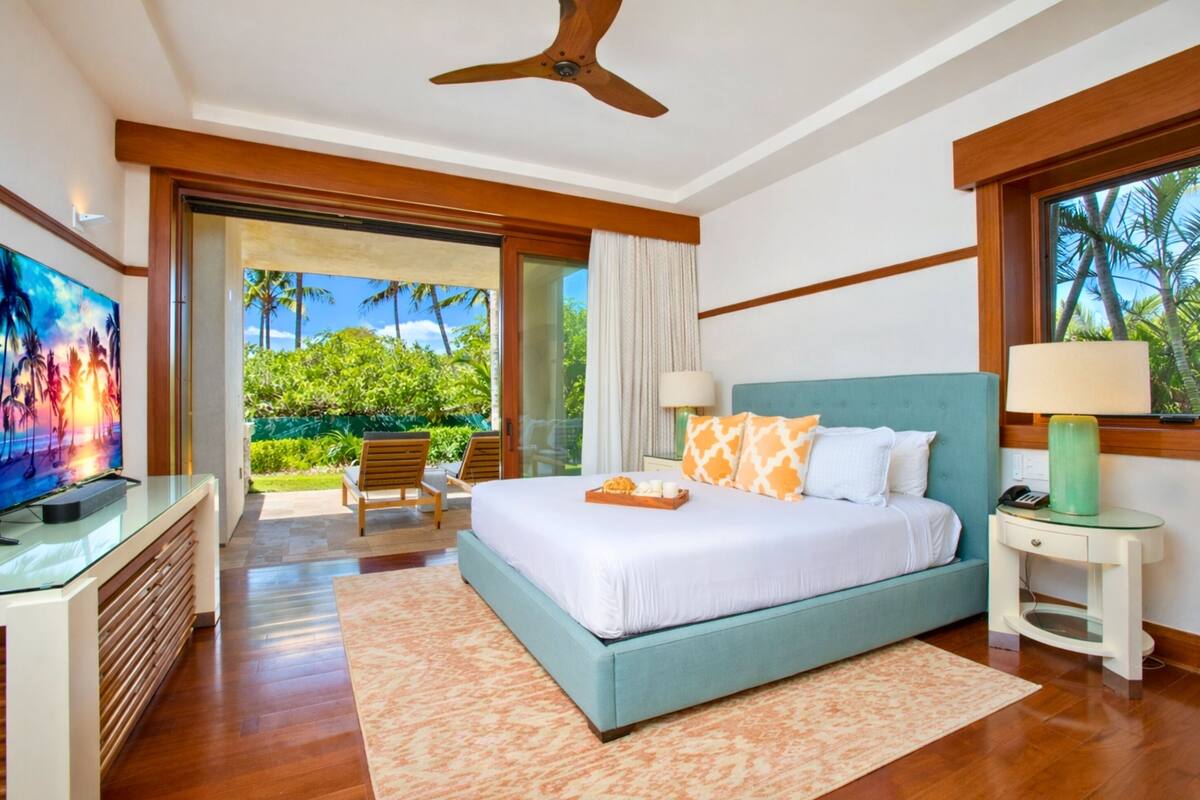 A bedroom with a bed, a TV, a ceiling fan, a nightstand, and a view of greenery outside.
