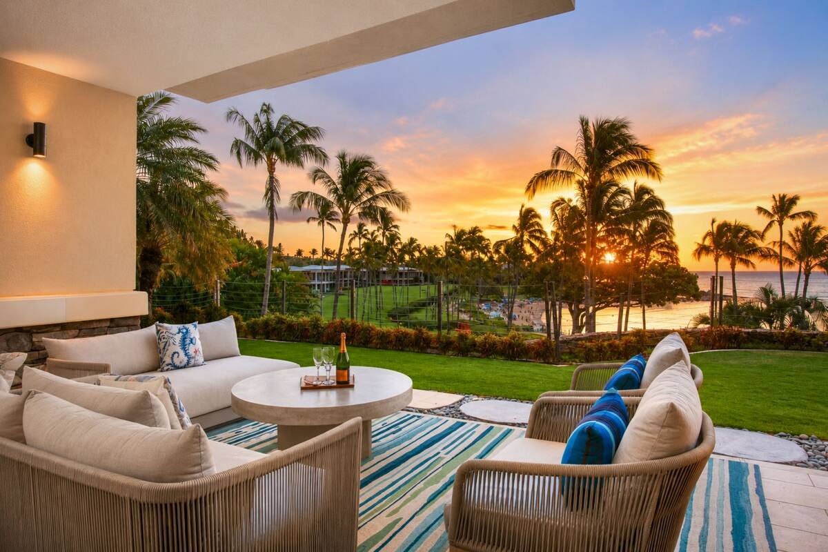 A patio with outdoor furniture, a table with a bottle and glasses, and a view of palm trees and the ocean at sunset.