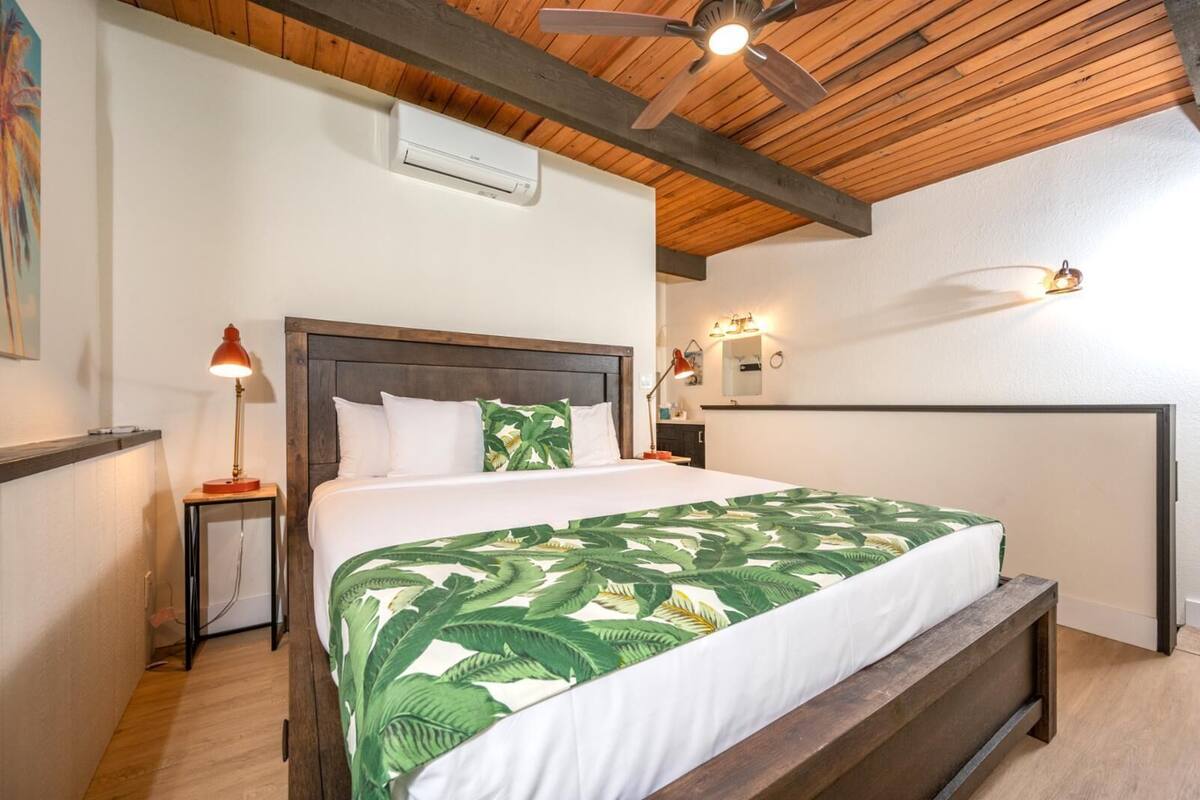 A bedroom with a wooden ceiling, a bed with a green leaf-patterned bedspread, a nightstand with a lamp, and a ceiling fan.