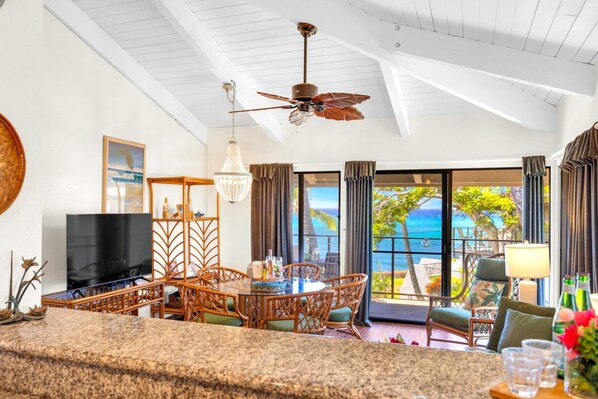 A spacious living area with a large TV, dining table, and chairs, a ceiling fan, and a view of the ocean. - Napili Point Resort by KBM (Napili-Honokowai)
