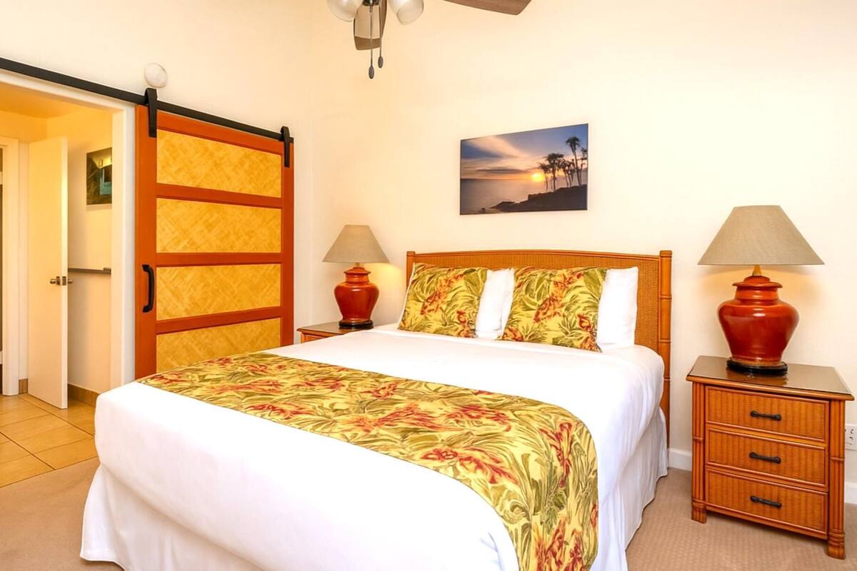 A hotel room with a bed, two bedside tables with lamps, a wooden door, a painting on the wall, and a ceiling fan.