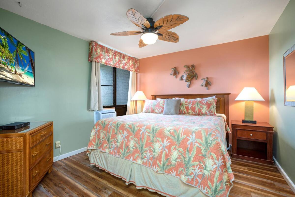 A bedroom with a bed, two bedside lamps, a ceiling fan, a window with curtains, a wall-mounted TV, and a wooden dresser.