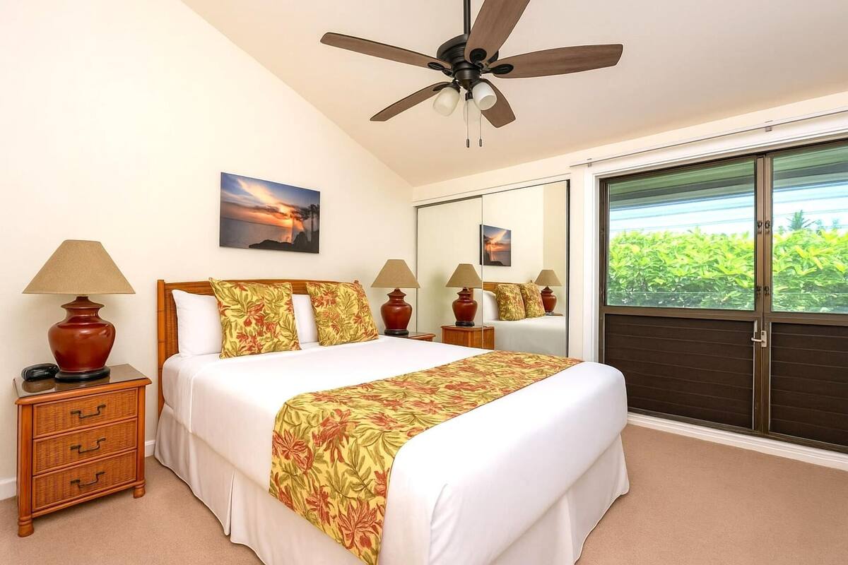 A hotel room with a bed, bedside lamps, a ceiling fan, a large window with a view of greenery, and a mirror.