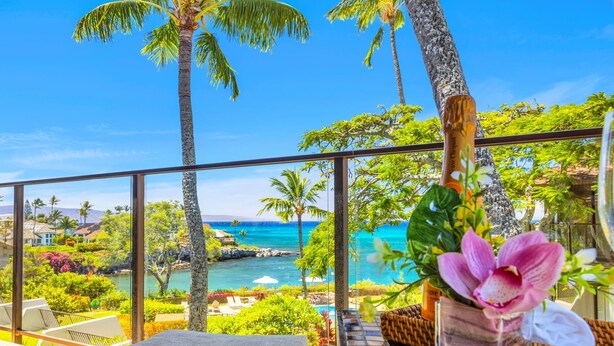Un balcón con vista a una playa y al océano, una silla de mimbre y un jarrón con flores.