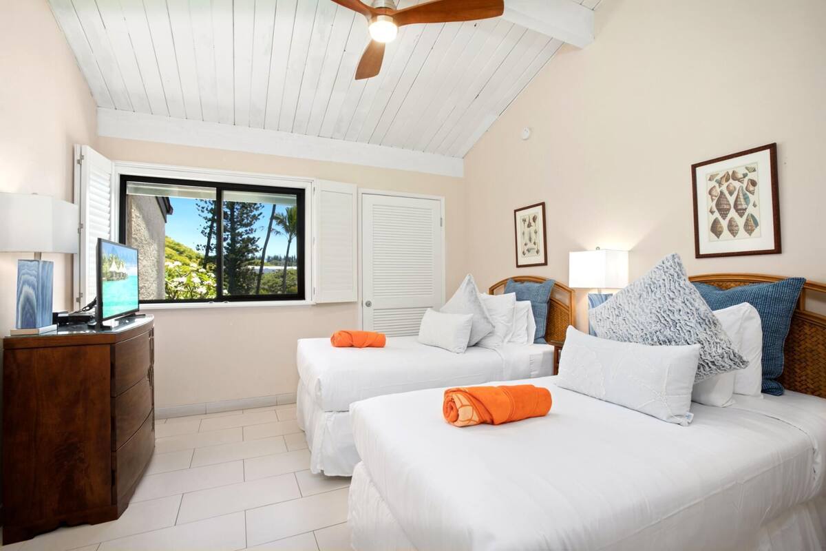 A hotel room with two beds, a desk with a computer, a window with a view of trees and a beach, and a ceiling fan.