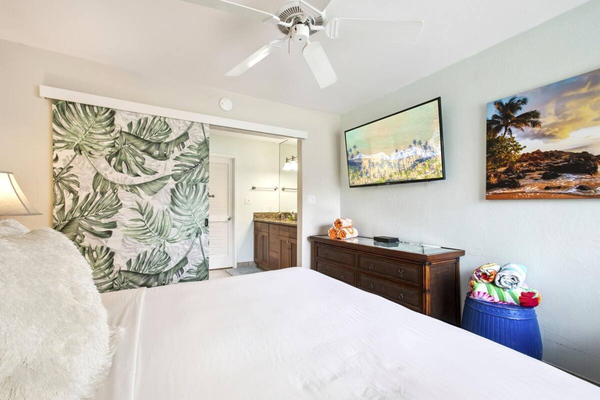 A bedroom with a bed, a ceiling fan, a TV, and artwork on the wall.