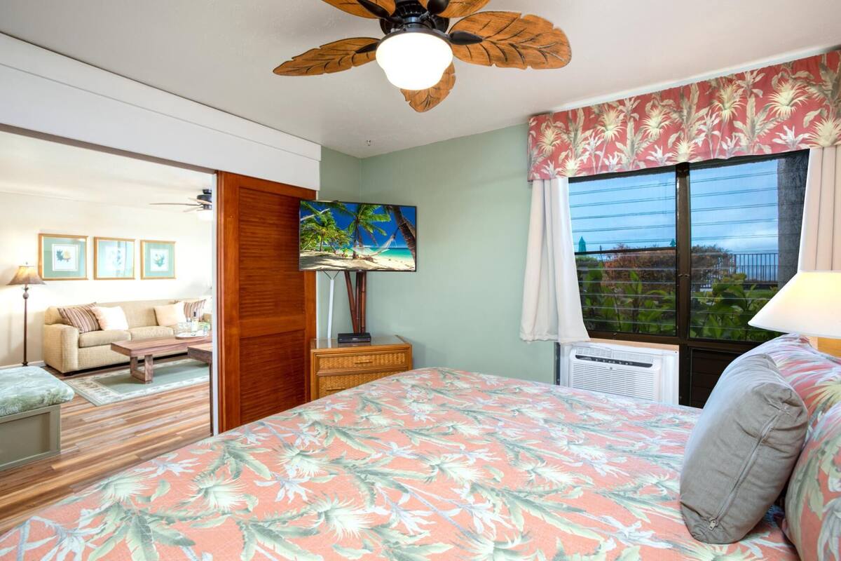 A bedroom with a bed, a TV, a ceiling fan, a window with curtains, and a view of the outdoors.