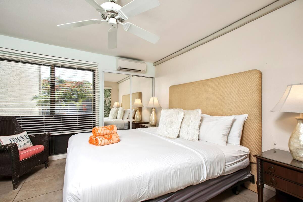 A hotel room with a bed, a ceiling fan, a window with blinds, a nightstand with a lamp, and a wicker chair.